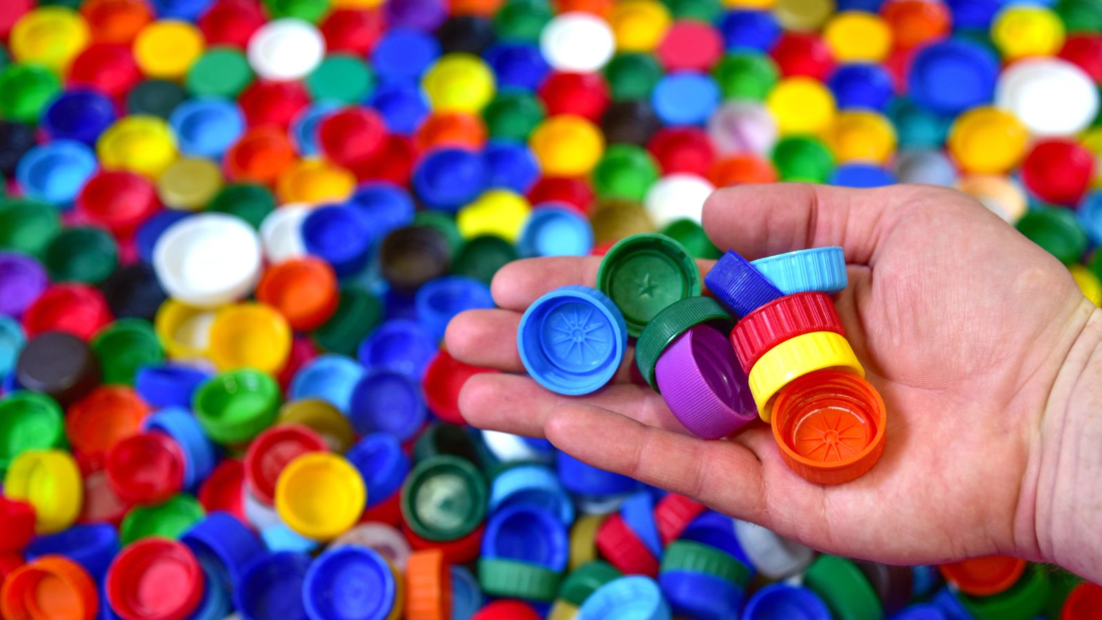 A close-up and overhead shot of a person in the process of holding a pile of bottle caps, with a large composition of the same material in the background