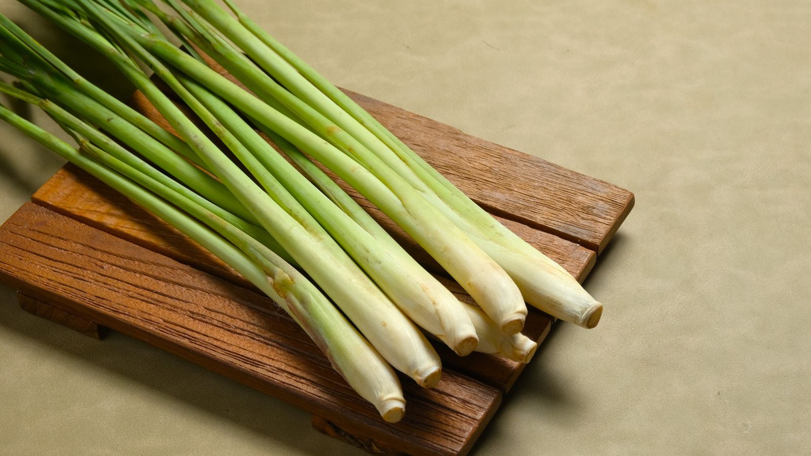 A wooden board appearing dark brown with multiple lemongrass pieces piled on top, appearing long with green stems that turn white on one end