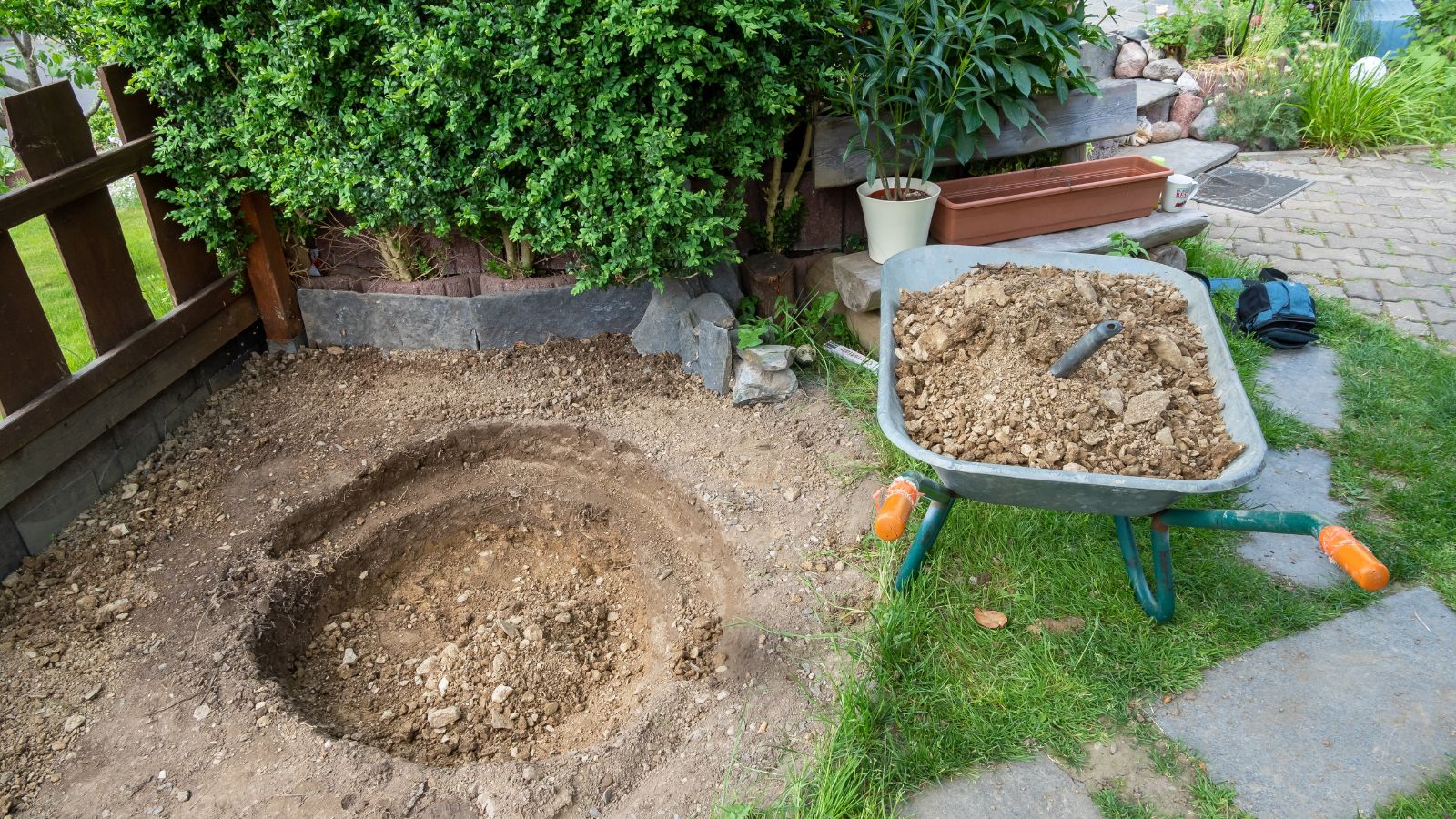 An overhead shot of a dug out hole beside a wheelbarrow, all situated in a yard area outdoors