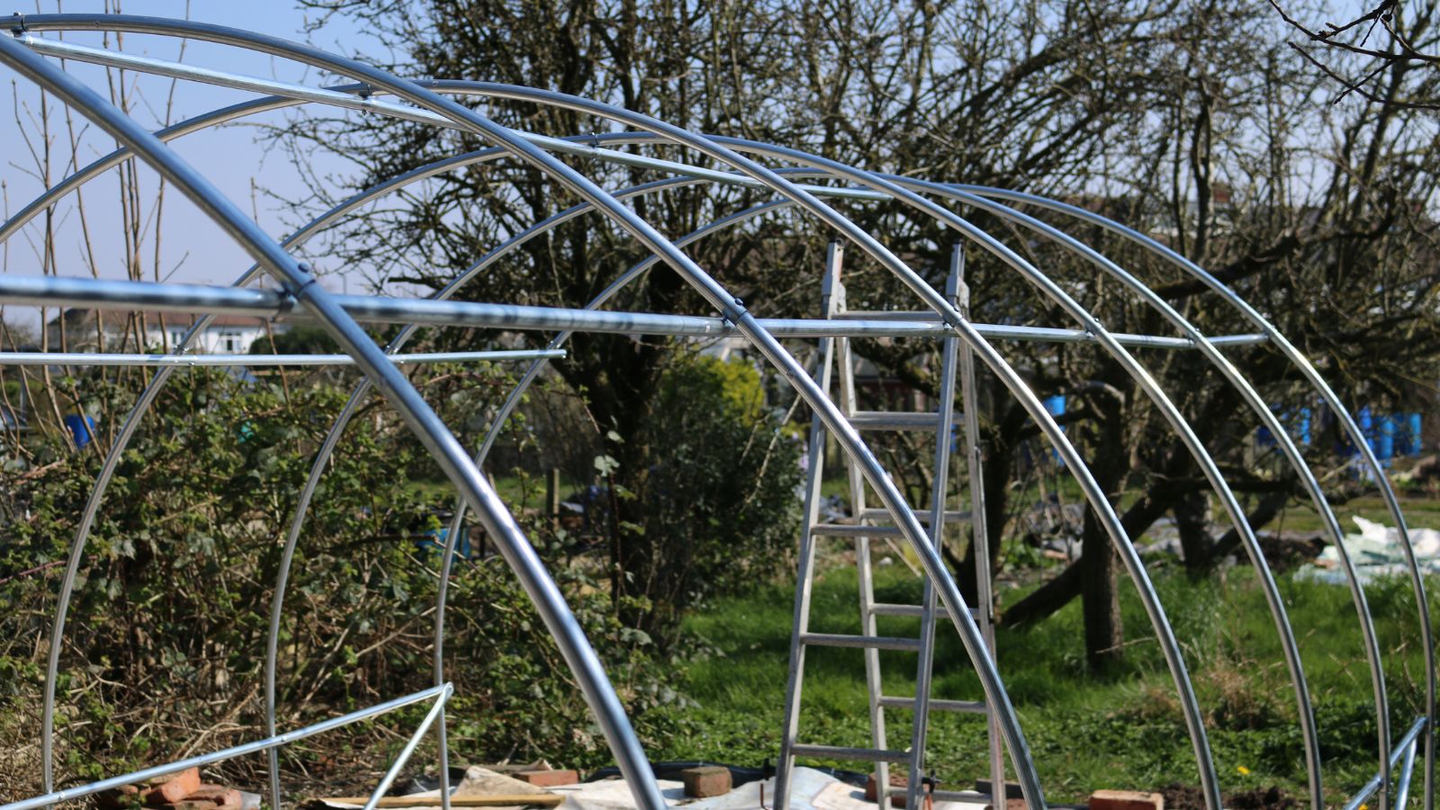 A frame meant to hold a plastic cover, with the structure made of steel poles and tubes with a ladder in the background