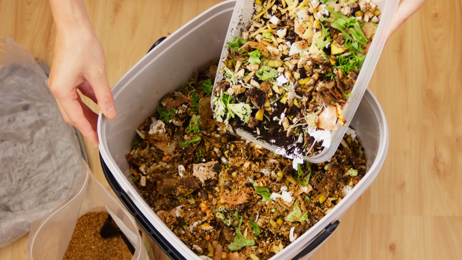 A person pours a mix of chopped vegetable scraps, peels, and leafy greens into a Bokashi composting bin, with a layer of brown, nutrient-rich material at the bottom.