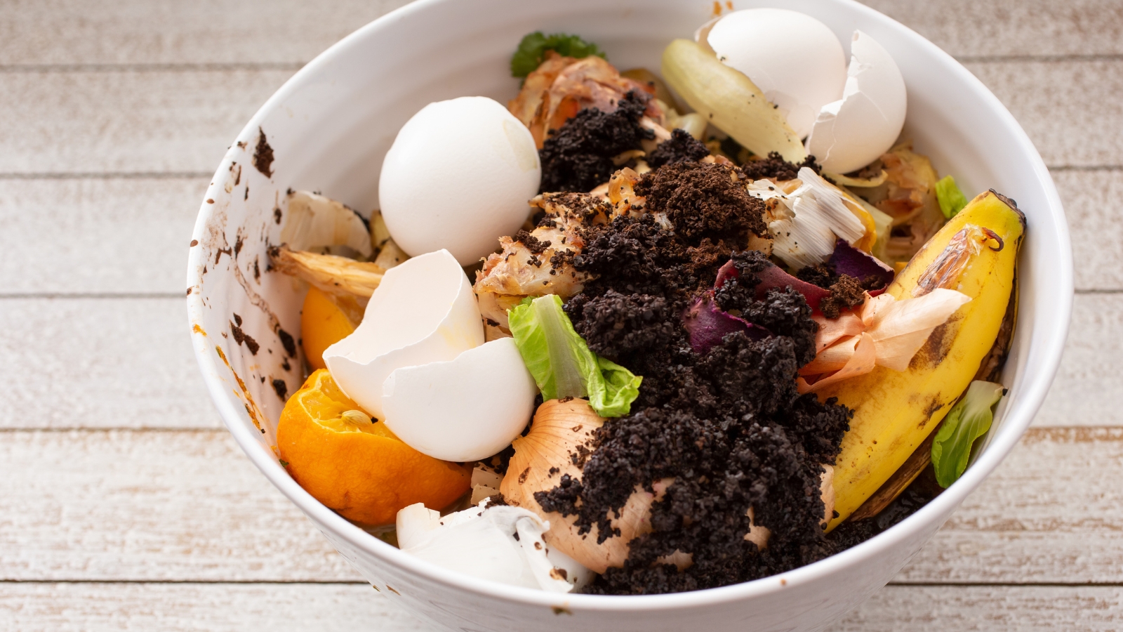 A white ceramic bowl filled with fruit peels, eggshells, and vegetable scraps is topped with a sprinkling of dark, moist organic material, set on a wooden surface.