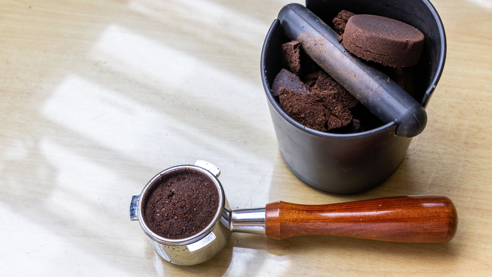 A metal tool with a wooden handle rests beside dark clumps of organic material inside a small black container, placed on a light-colored wooden table.