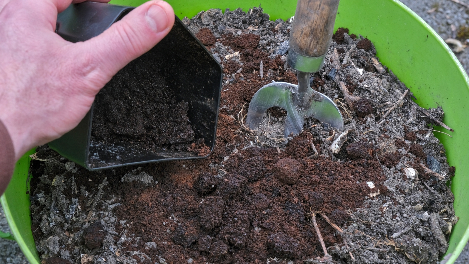 A black scoop transfers damp, crumbly organic matter into a large green container, where it is surrounded by dry soil and small twigs.