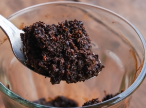 A metal spoon holds a clump of damp, dark brown organic matter with a granular texture, resting above a transparent glass bowl on a wooden surface.