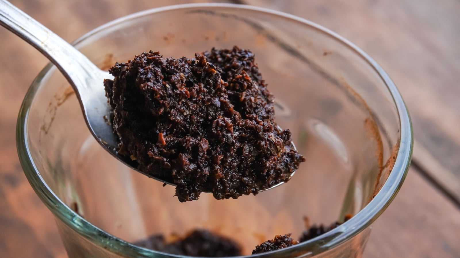 A metal spoon holds a clump of damp, dark brown organic matter with a granular texture, resting above a transparent glass bowl on a wooden surface.