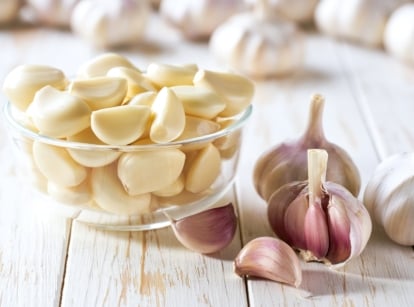 A small glass bowl filled with pale white, crescent-shaped cloves sits on a light wooden surface, surrounded by larger unpeeled bulbs with papery skins in shades of purple and white.