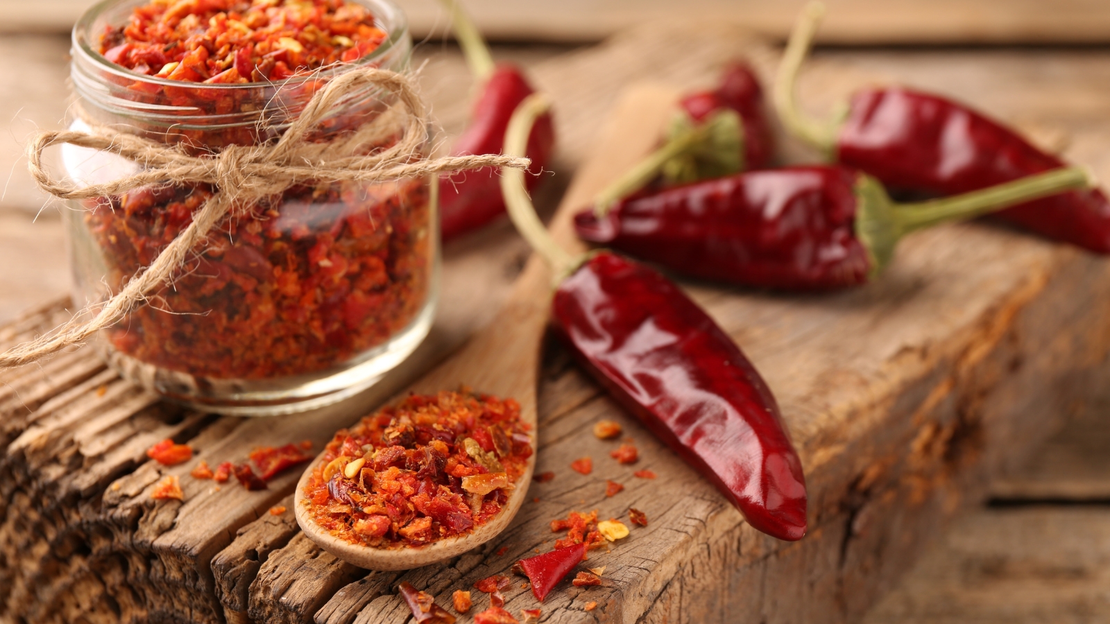 A glass jar filled with bright red ground spice sits next to dried red peppers, some tied with twine, scattered on a rough wooden surface with visible cracks and textures.