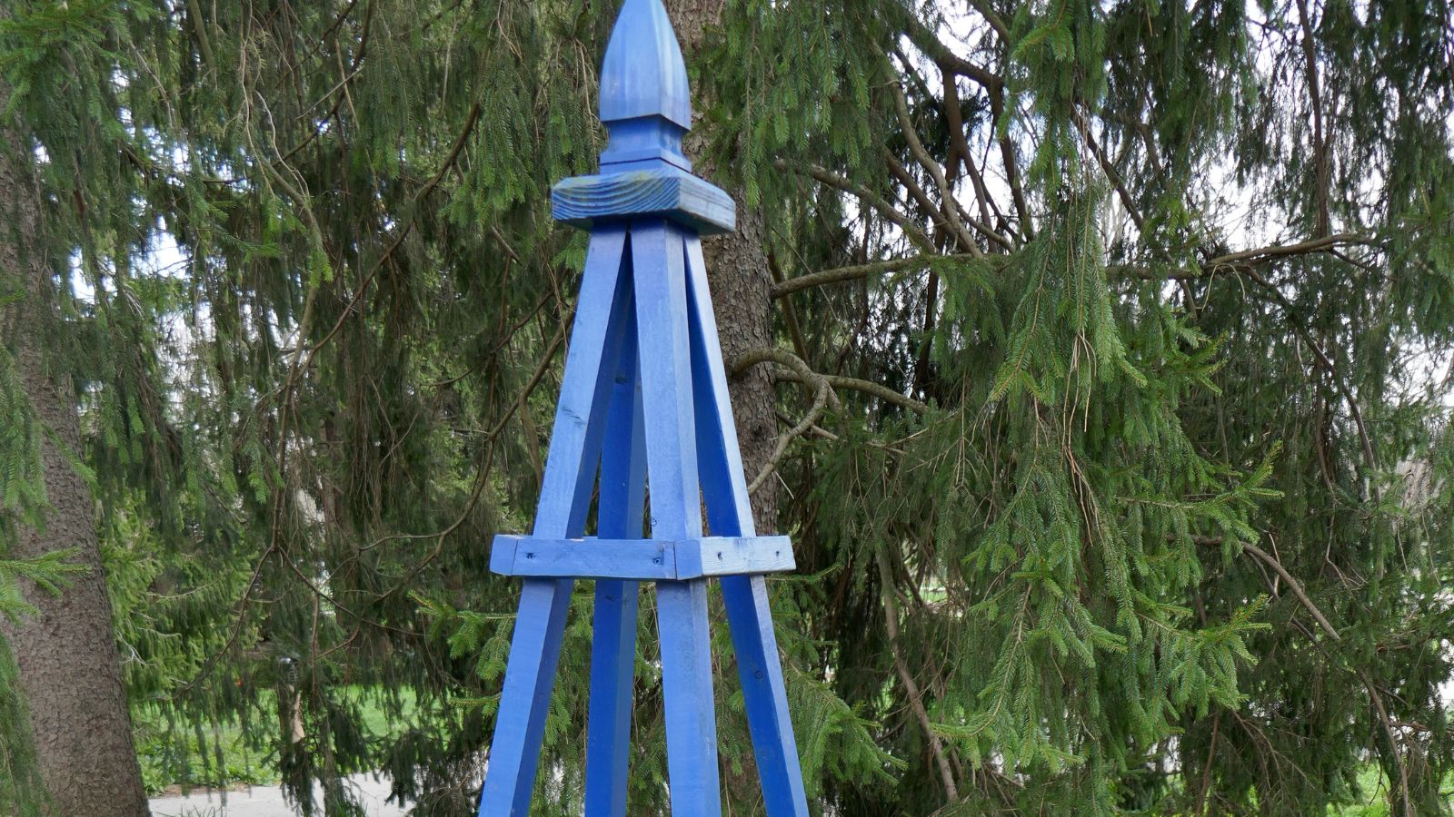 A close up of the top of a pointed tomato cage painted a bright blue with many trees in the background
