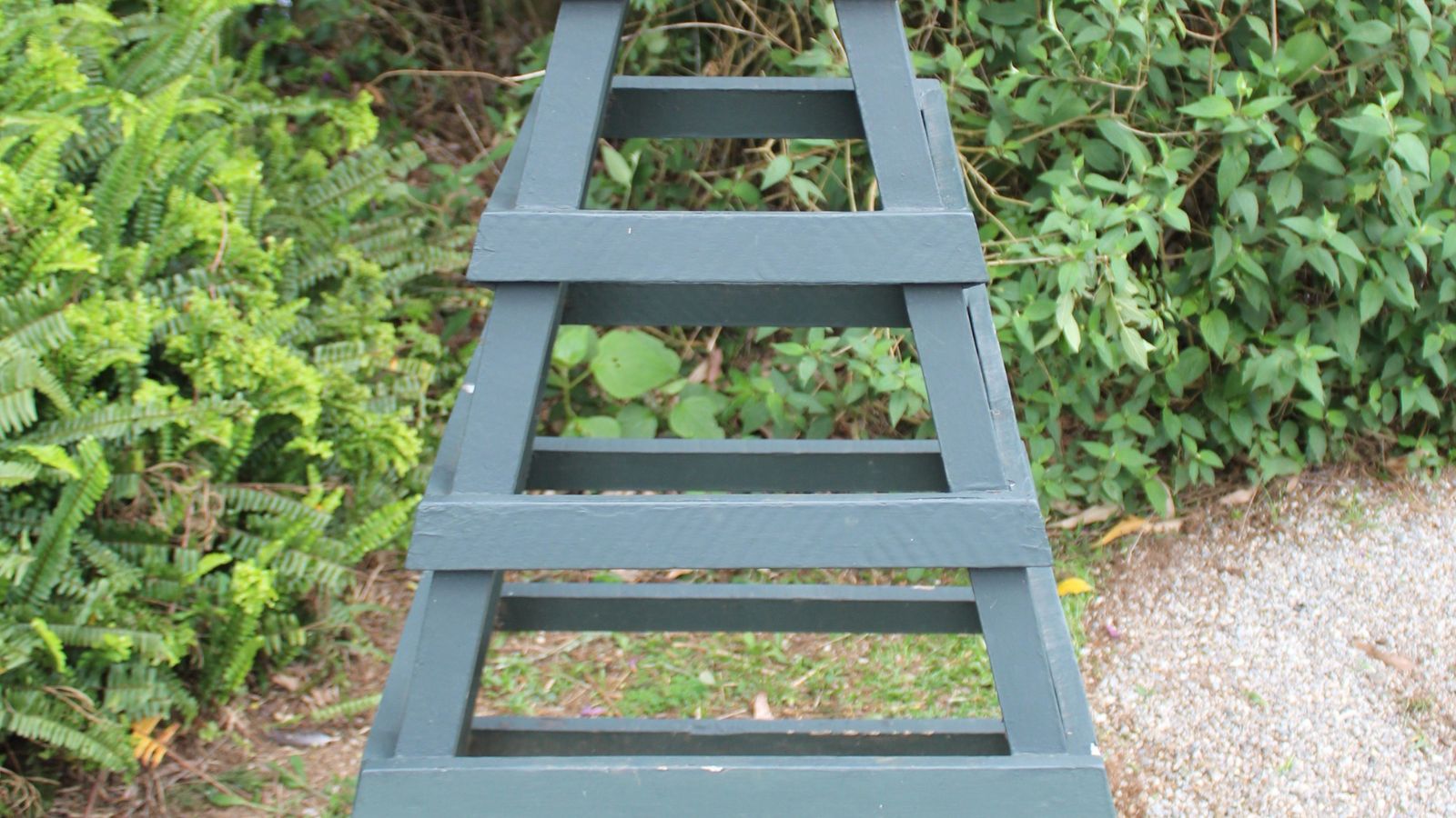 A wooden garden obelisk appearing sturdy and painted with a deep bluish gray color with other foliage in the background