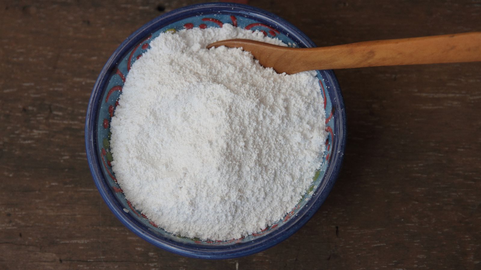 A close-up and overhead shot of a small bowl filled with Kaolin Clay powder, all placed on a wooden surface in a well lit area indoors