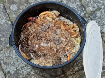 A close-up and overhead shot of a black colored bucket, filled with bran and food scraps, showcasing how to make a DIY bokashi