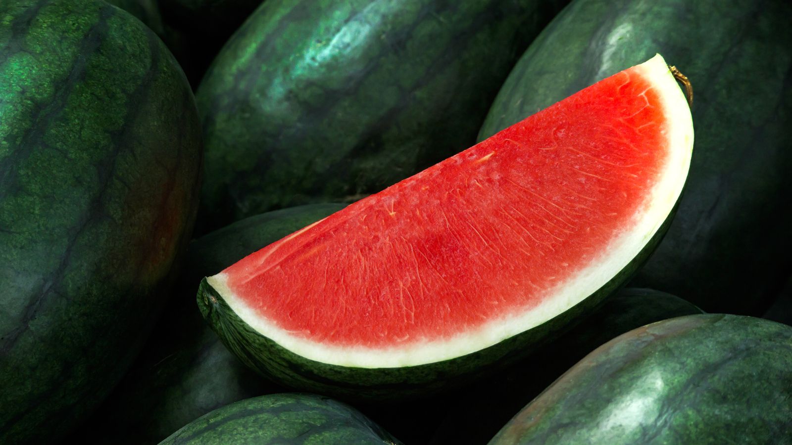 A slice of Seedless Watermelon appearing to have pure red flesh placed on top of a pile of other unsliced crops with deep green color