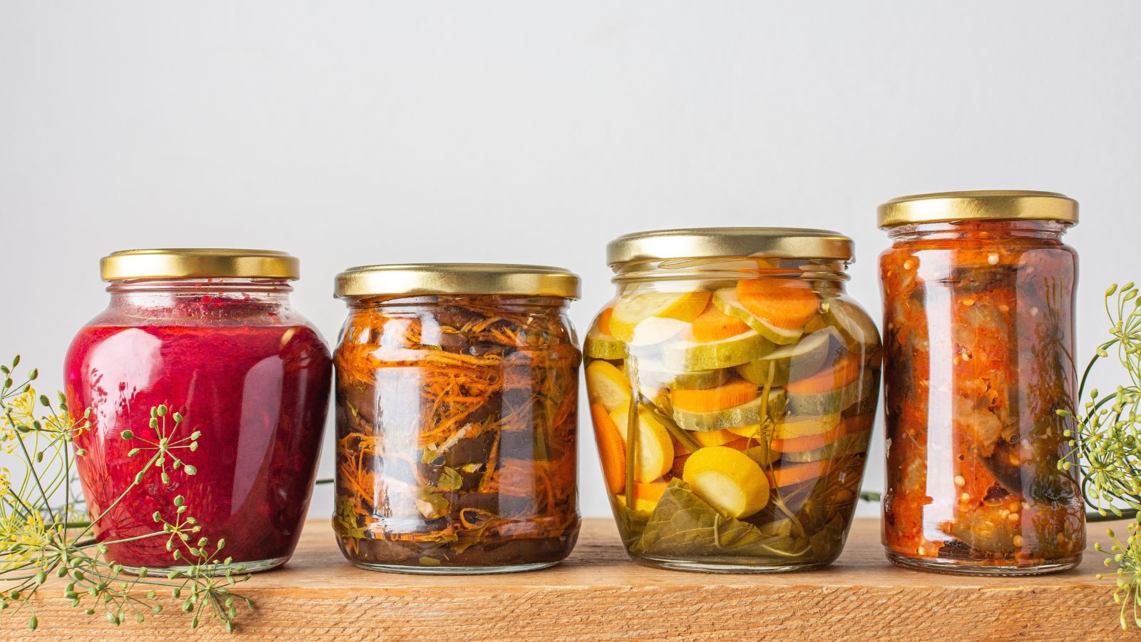 Glass jars filled with fermenting vegetables placed in a row on a wooden surface, with lovely thin stems placed near the base