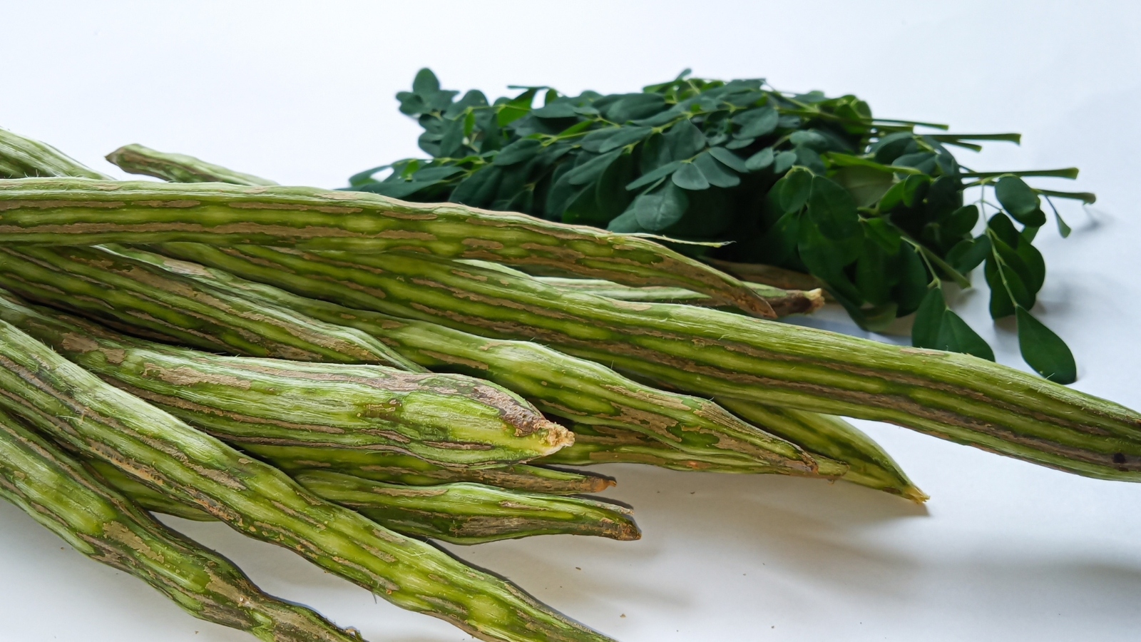 A fresh bunch of long, thin, green seed pods with smooth skin, lying on a white surface alongside leafy branches, ready for harvest.