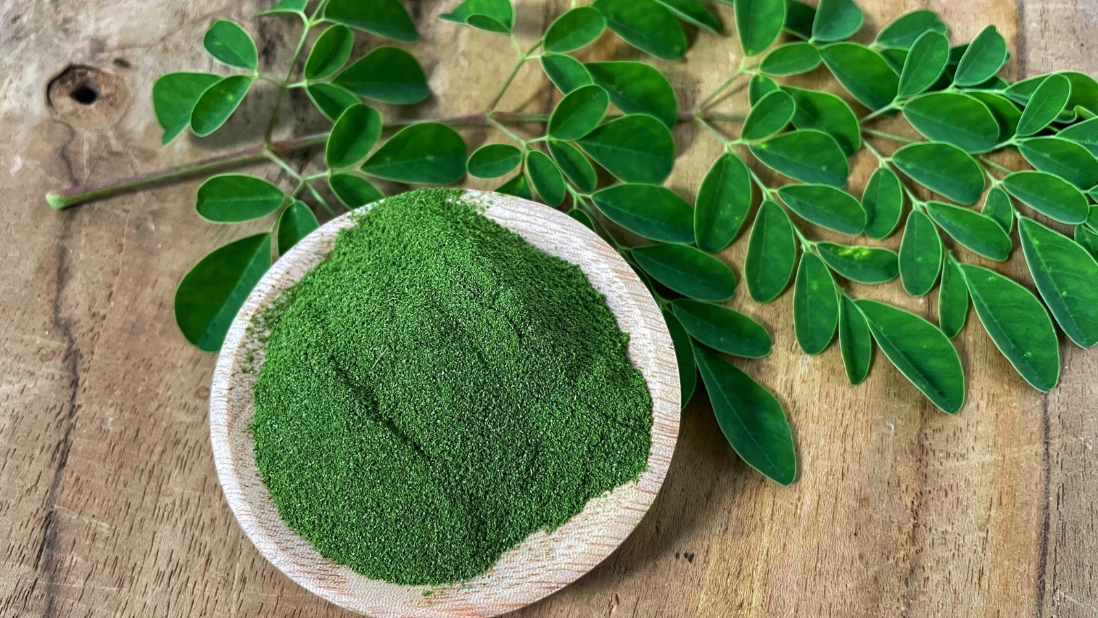 A bowl of vibrant green powder sits on a rustic wooden surface, surrounded by fresh green leaves arranged neatly around the bowl.