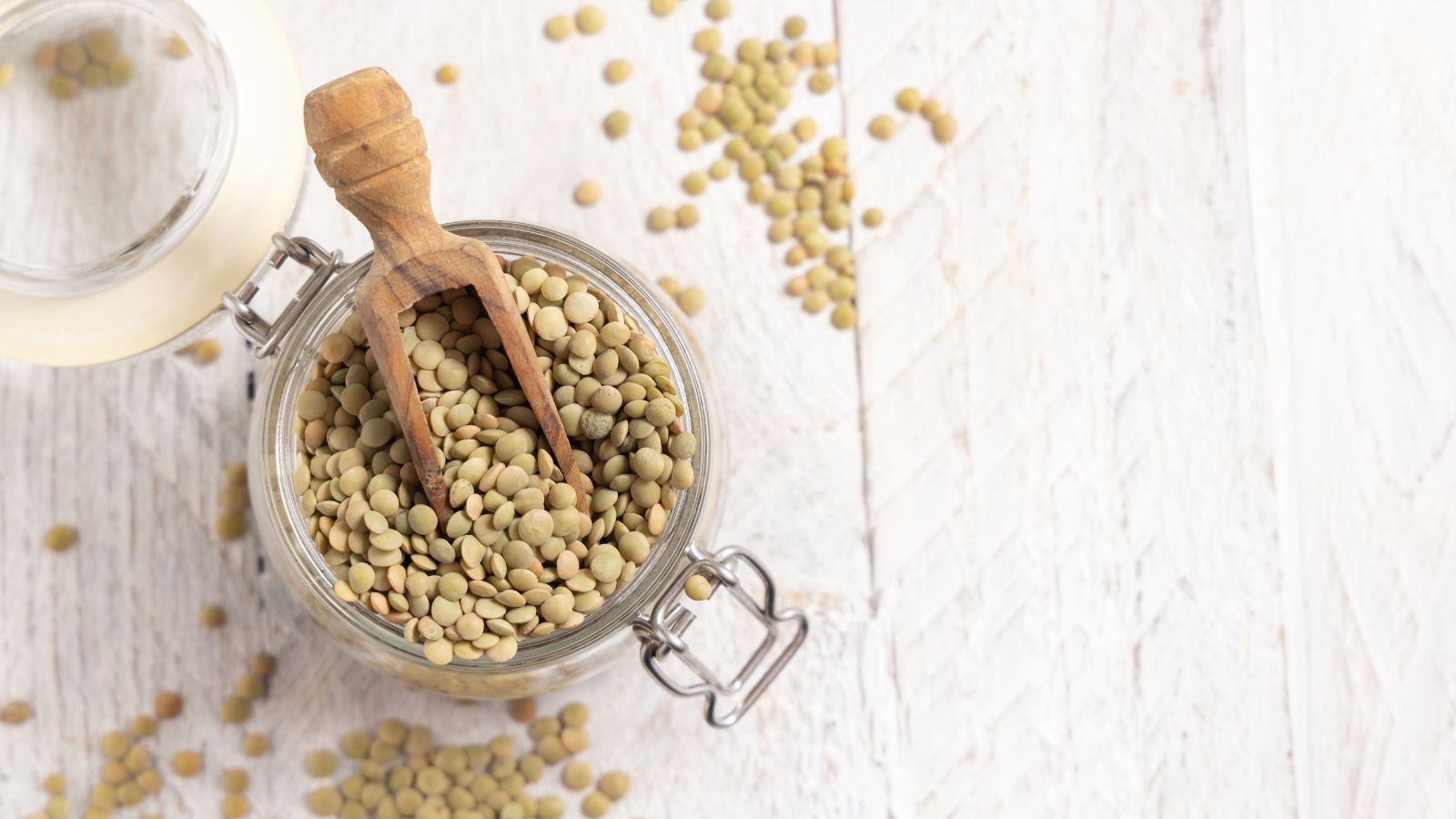 Many Lens culinaris seeds in a jar with a wooden scooper placed into the seeds placed on a white wooden surface