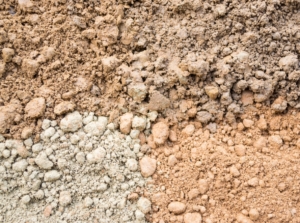 A close-up of a dry, uneven ground surface featuring a mix of light brown and pale grey textures, marked with fine cracks and small clumps.