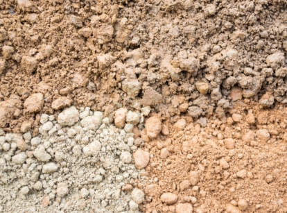 A close-up of a dry, uneven ground surface featuring a mix of light brown and pale grey textures, marked with fine cracks and small clumps.