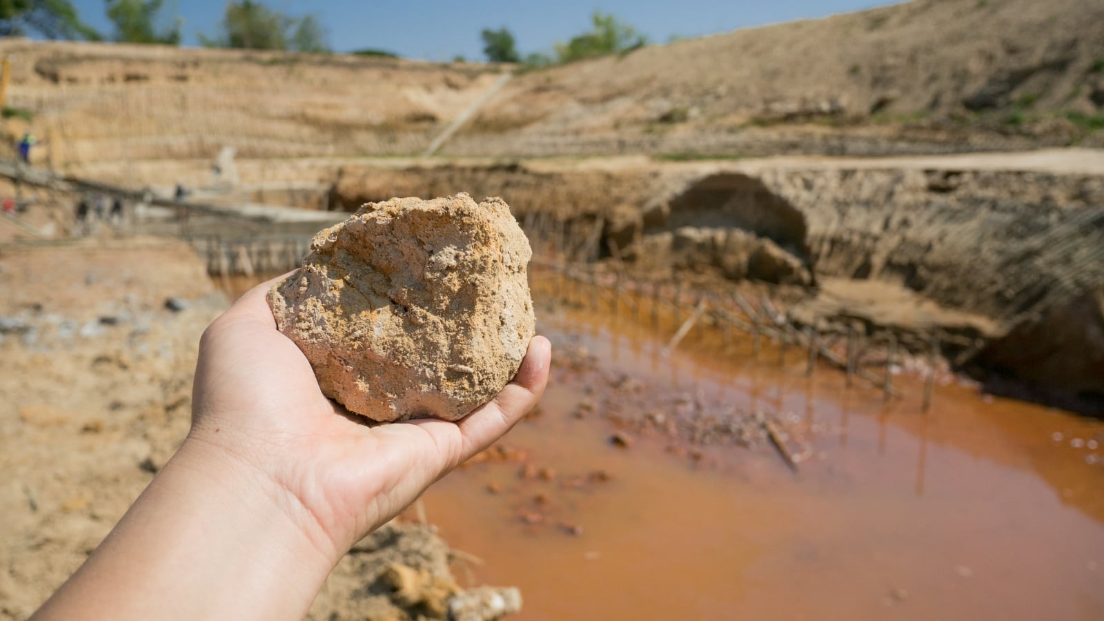 A hand holding up a sizable, jagged rock over an orange-tinted water channel, where muddy, cracked banks meet standing water.
