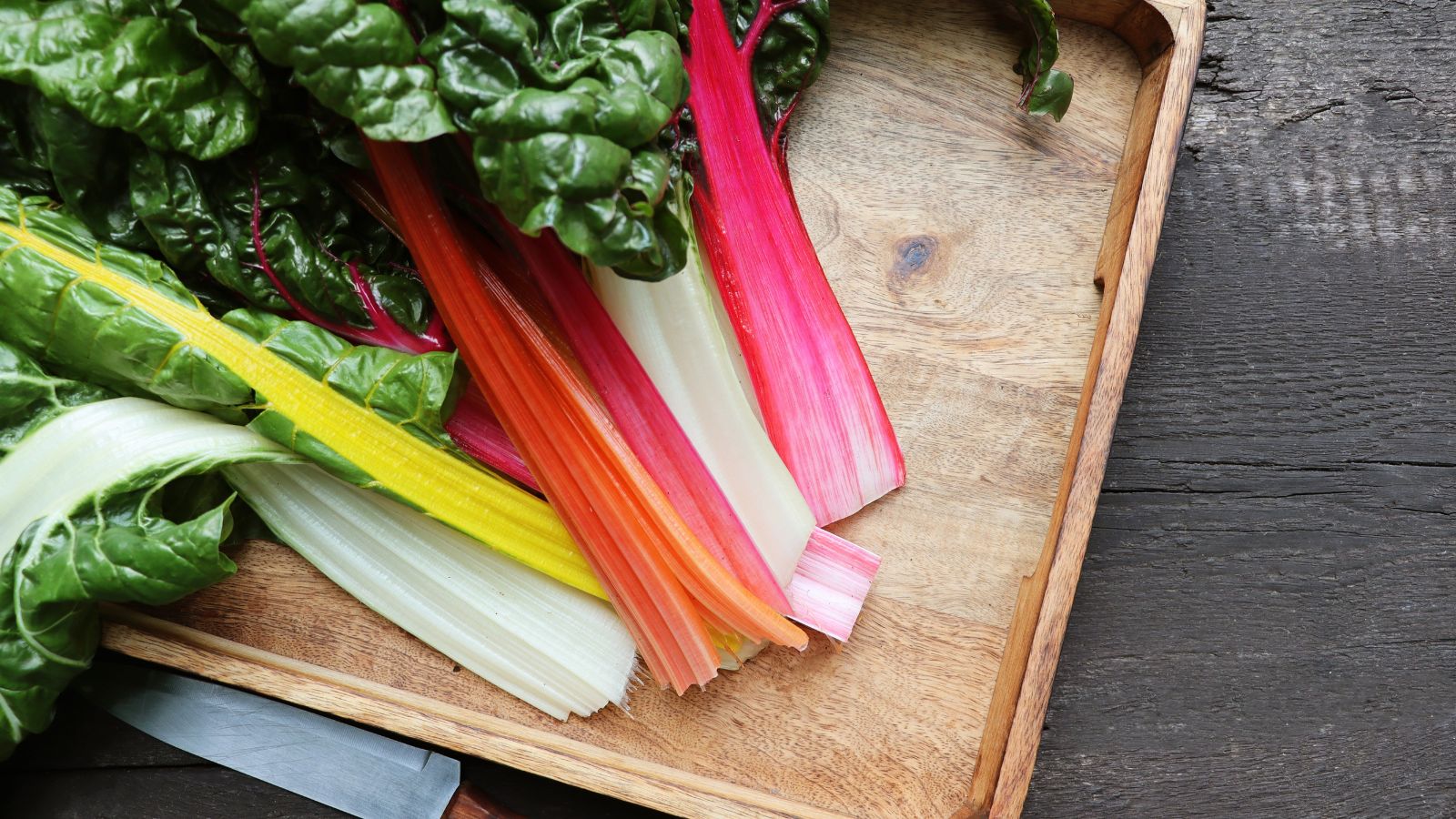 A close-up and overhead shot of a composition of freshly harvested leafy greens, all placed on a wooden tray beside a knife, all situated in a well lit area indoors