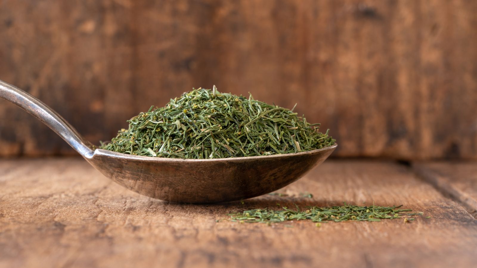A spoonful of dried herbs placed on a brown surface with a woody brown wall in the background
