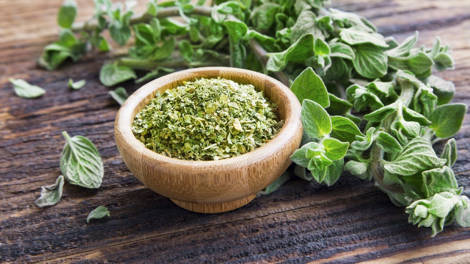 A close up shot of fresh and dehydrated herbs, placed on a small wooden bowl alongside freshly picked leaves, all situated in a well lit area