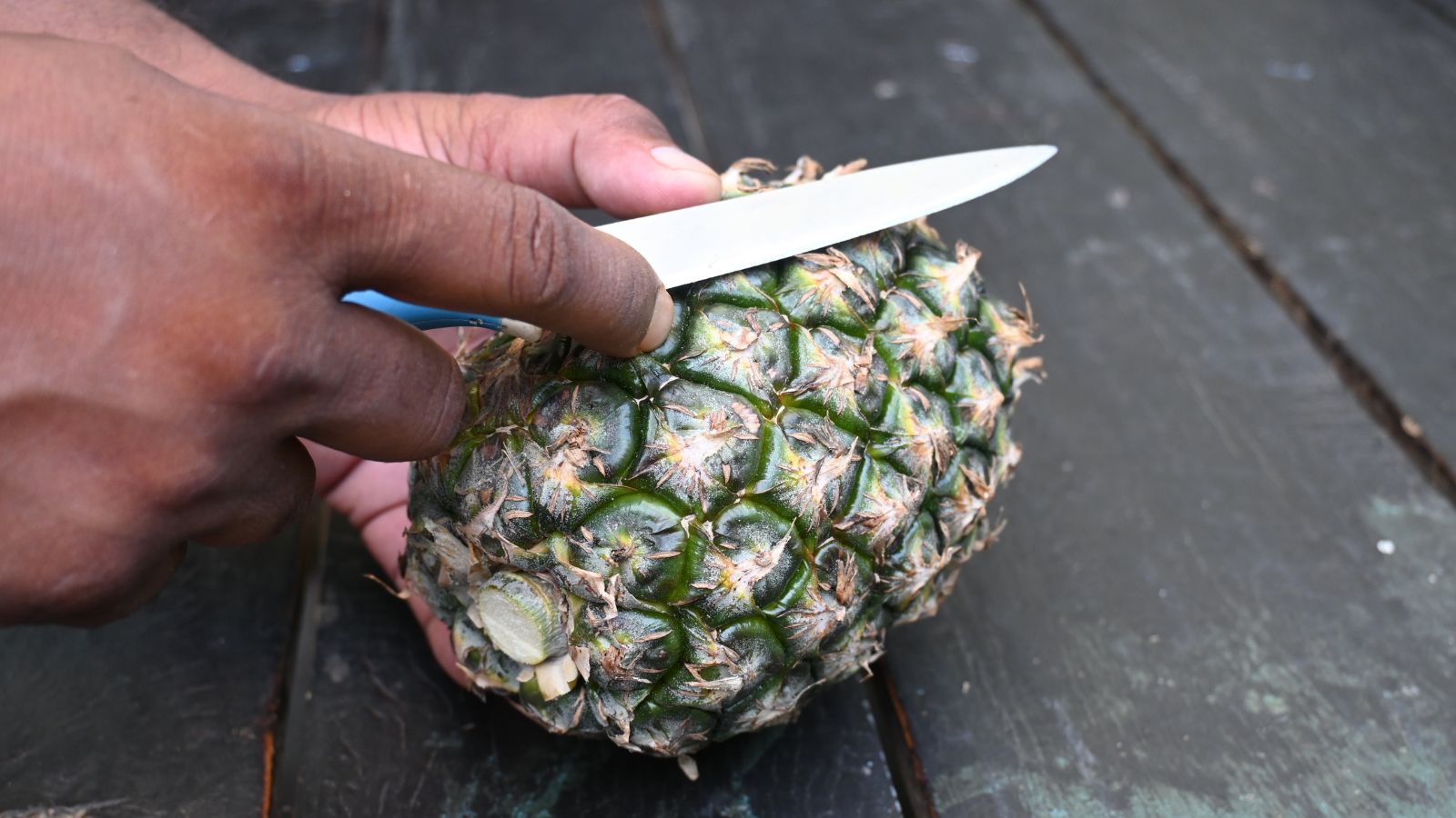 A person peeling a Ananas comosus fruit, with the top spiky part removed and then set on a wooden surface