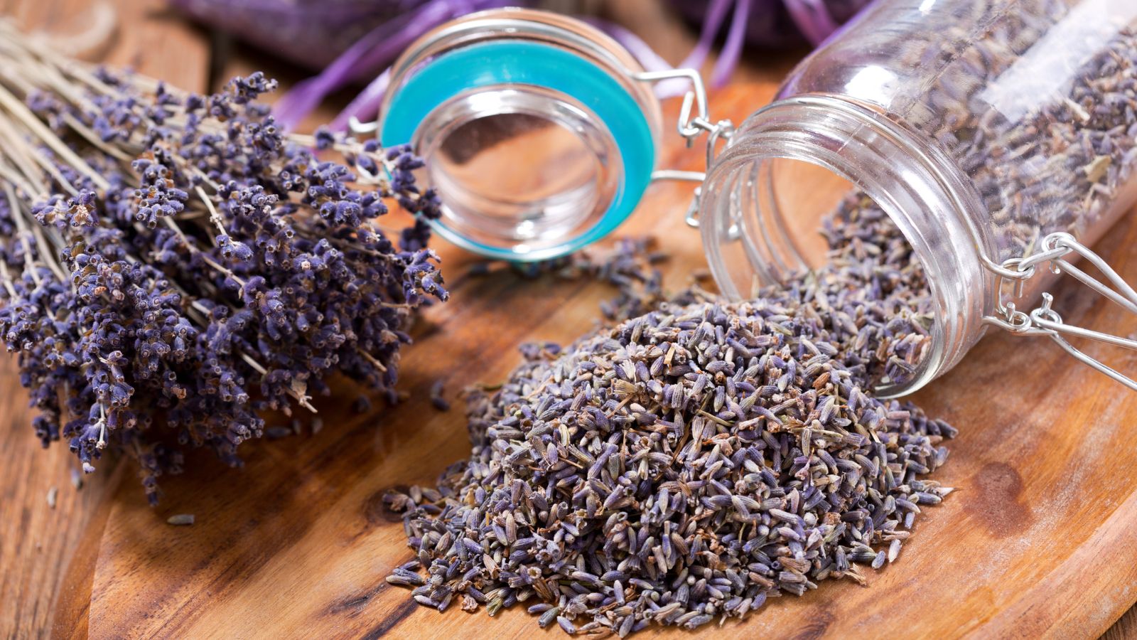 A close-up shot of dried purple flowers in a jar alongside the same flowers and its stems, all placed on a wooden surface in a well lit area