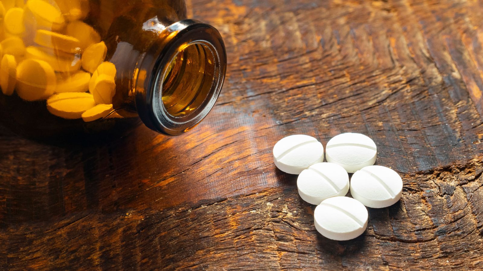 An amber bottle fallen over to reveal five white tablets on the wooden surface, with some tablets still inside the bottle