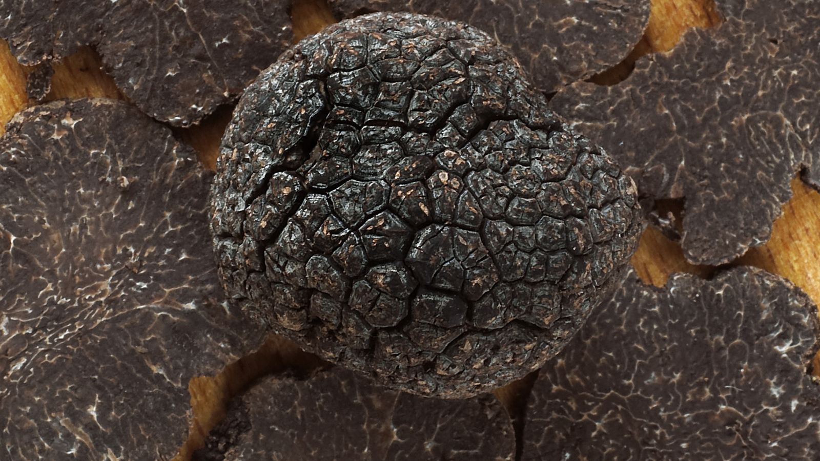An intact piece of Tuber melanosporum placed on a wooden surface with scattered slices of Tuber melanosporum
