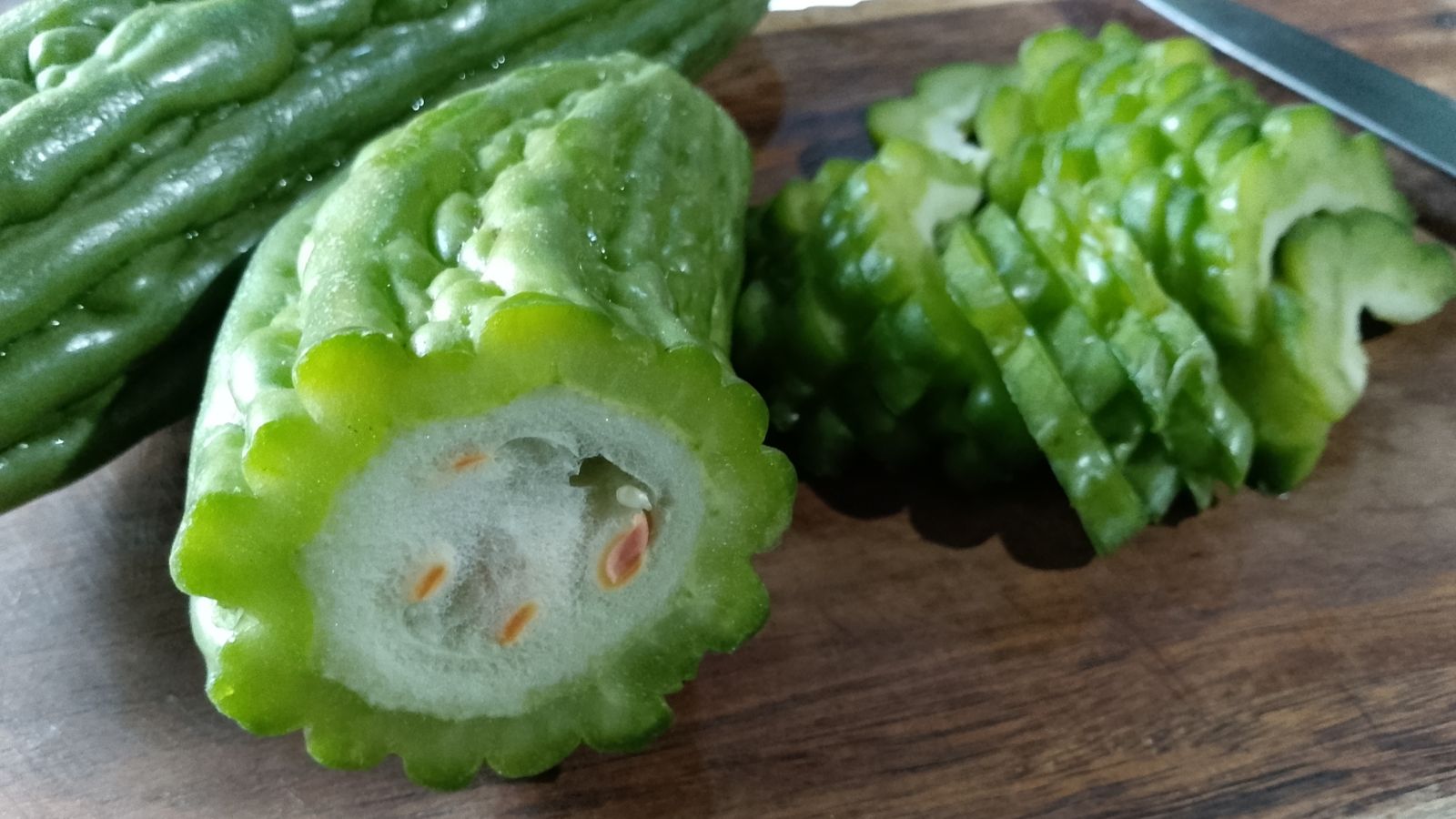 A sliced Momordica charantia crop exposing its white interior with tan seeds, appearing to have green bumpy skin placed on a wooden cutting board